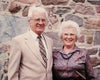 Paul and Edna stand in front of a field stone wall, smiling together.