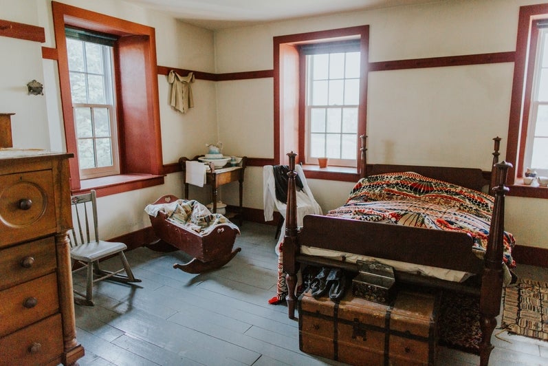 A small bedroom with three large windows, dresser, baby's crib, and bed with a quilt