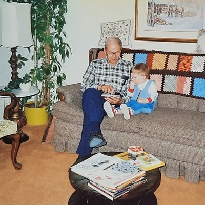 On a grey couch in a living room, Howard sits with a young child reading a cardboard book.