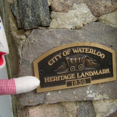 A hand puppet, featuring overalls and a mustache, points to a plaque that reads "City of Waterloo Heritage Landmark, 1850"