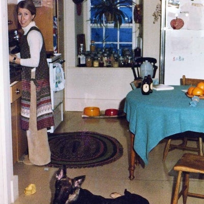 a young woman smiles in this vintage photo, while she stands at the kitchen counter. A young german shepherd dog lays on the kitchen linoleum floor.