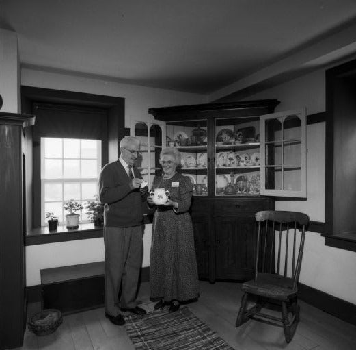 A black and white photo of Paul and Edna Hunsberger hold a tea set in Brubacher House, standing beside a cabinet with dishes and tea cups. 