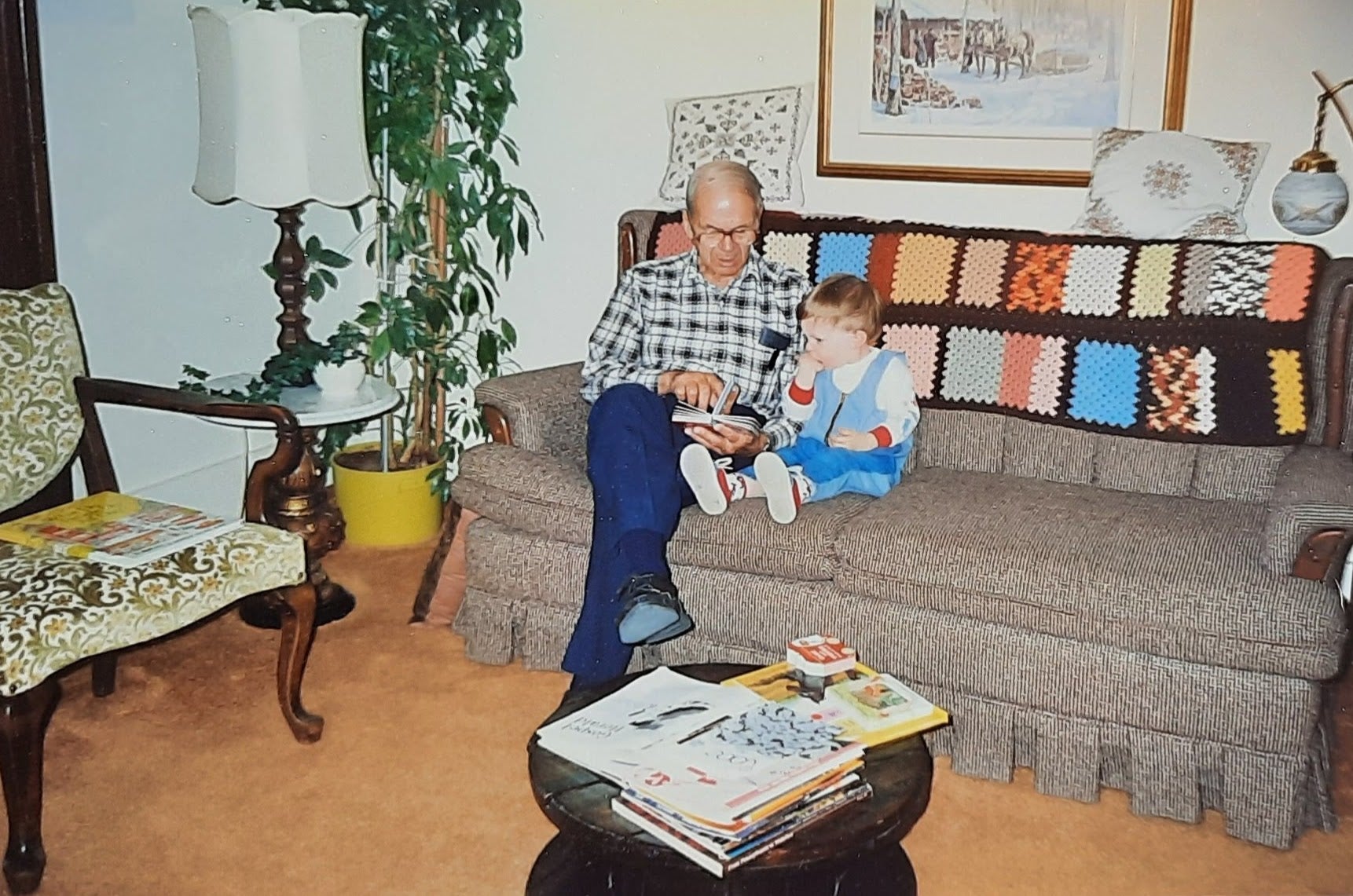 On a grey couch in a living room, Howard sits with a young child reading a cardboard book.