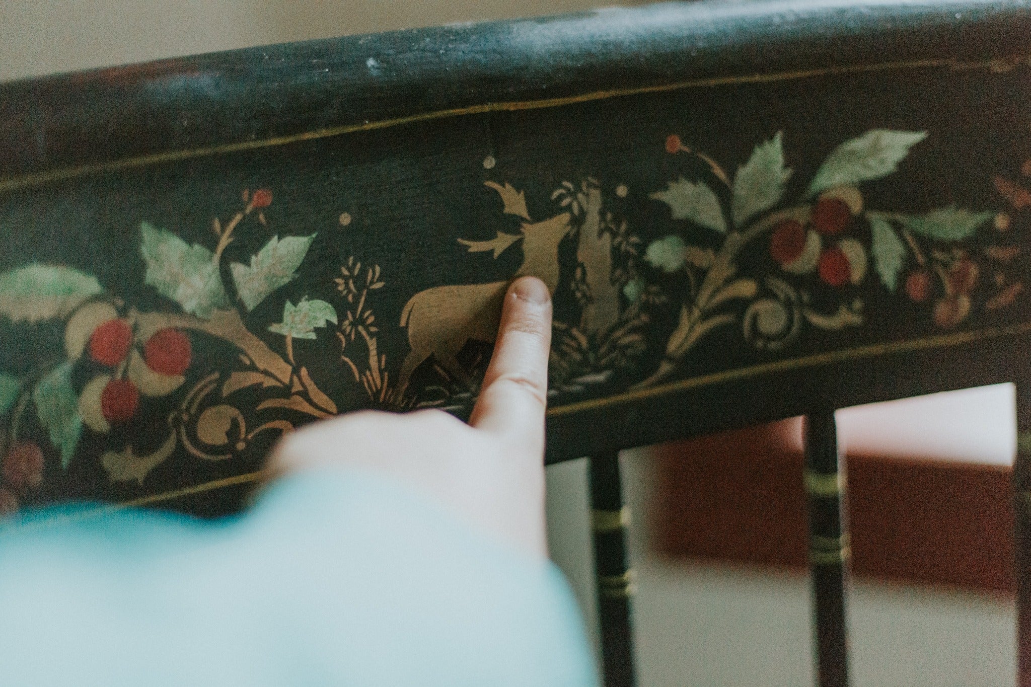 A finger points to a hand painted back of a chair, featuring a golden deer, some light green leaves and red raspberries