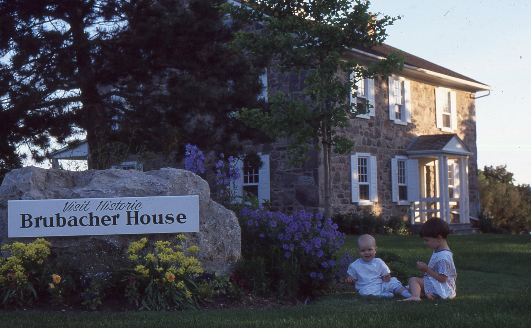 Two small children sit in the grass outside Brubacher House, beside the rock sign with flowers