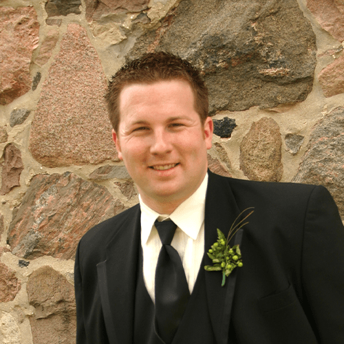 Chris, wearing a tux, stands in front of a field stone wall