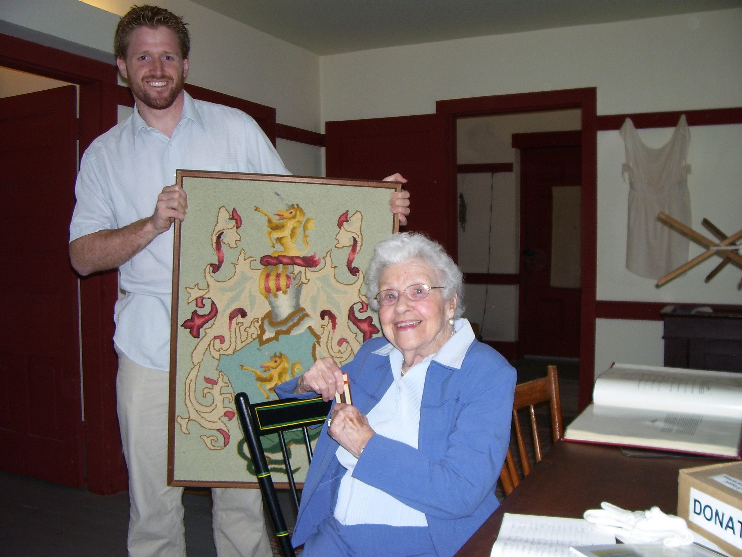 Brandon holds a textile with the Brubacher family crest. Dorothy Schweitzer Elliot, an elderly woman, sits in front.