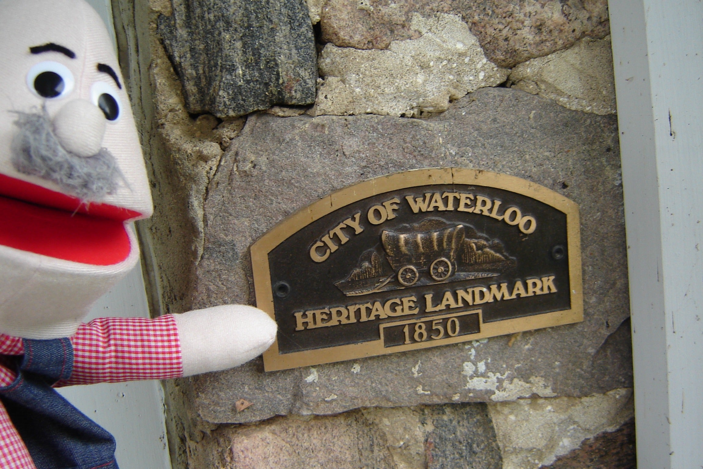A hand puppet, featuring overalls and a mustache, points to a plaque that reads "City of Waterloo Heritage Landmark, 1850"