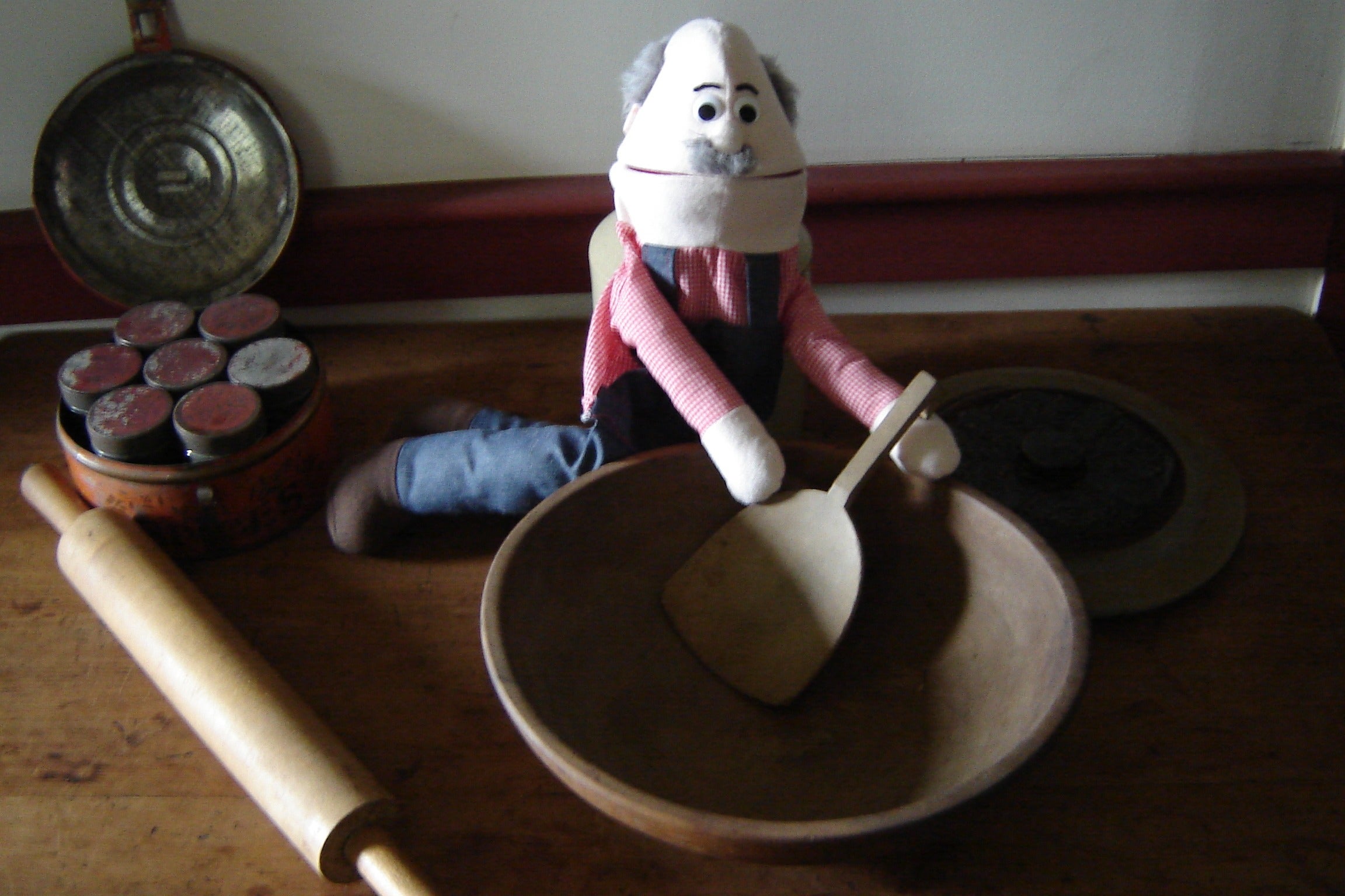 A hand puppet, featuring overalls and a mustache, sits on a table holding a spoon in a wooden bowl