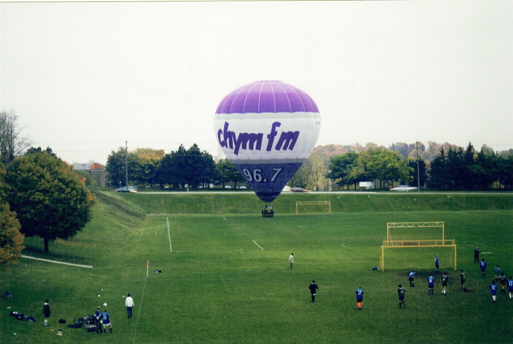 A purple and white hot air balloon takes off from a field. a few people are gathered around it.