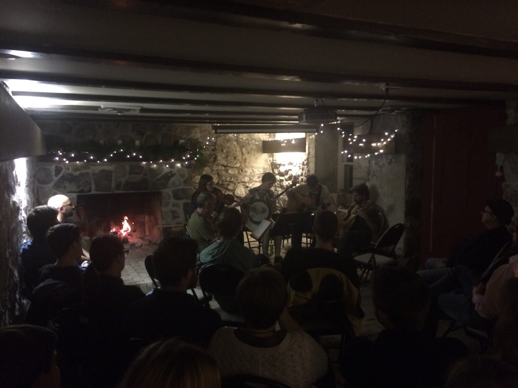 A darkly lit basement, with lights and a fire in the fireplace, a folk band plays in the corner, enjoyed by a crowd.