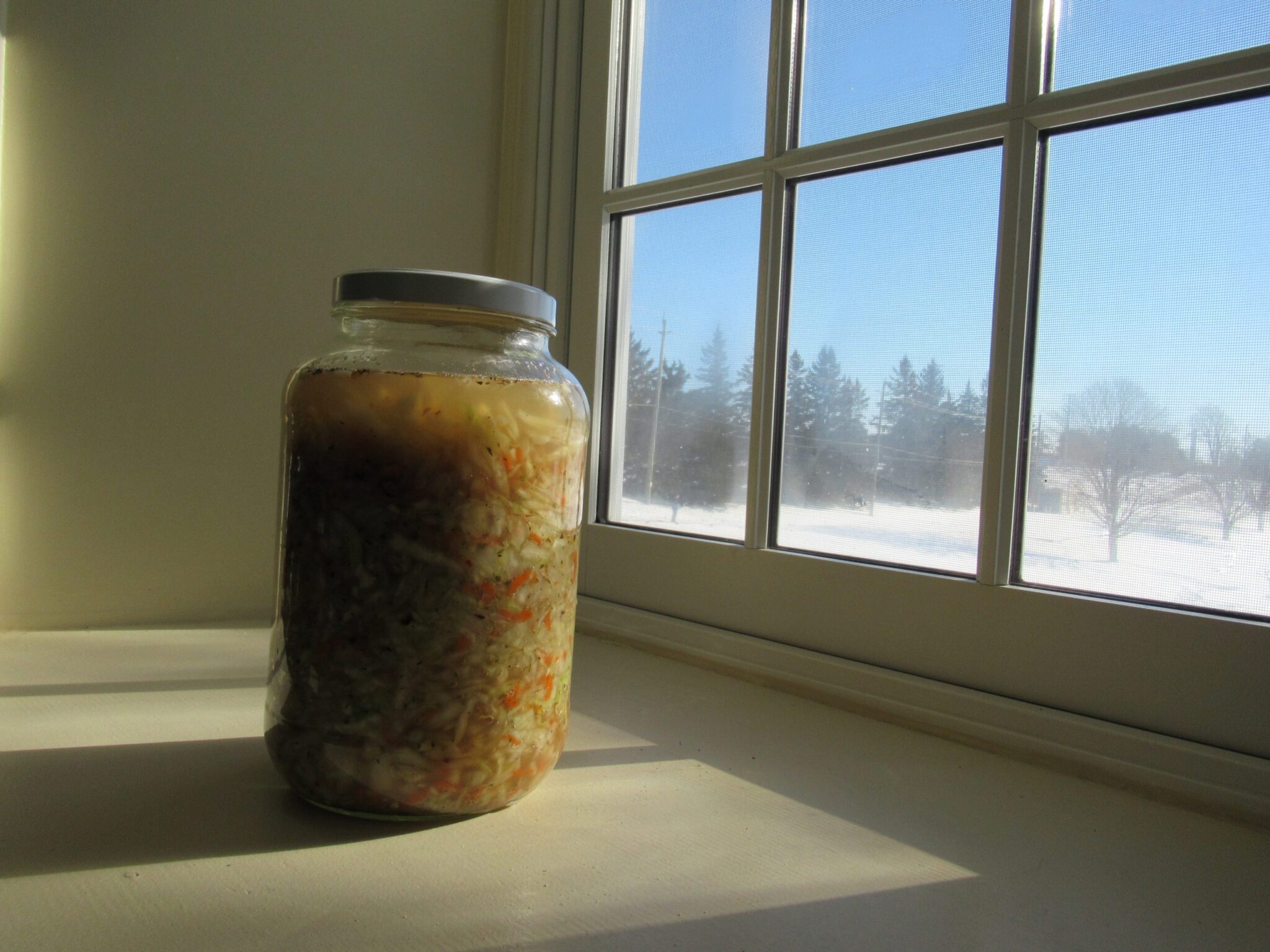 A mason jar of sauerkraut sits on the window sill, a winter scene outside.