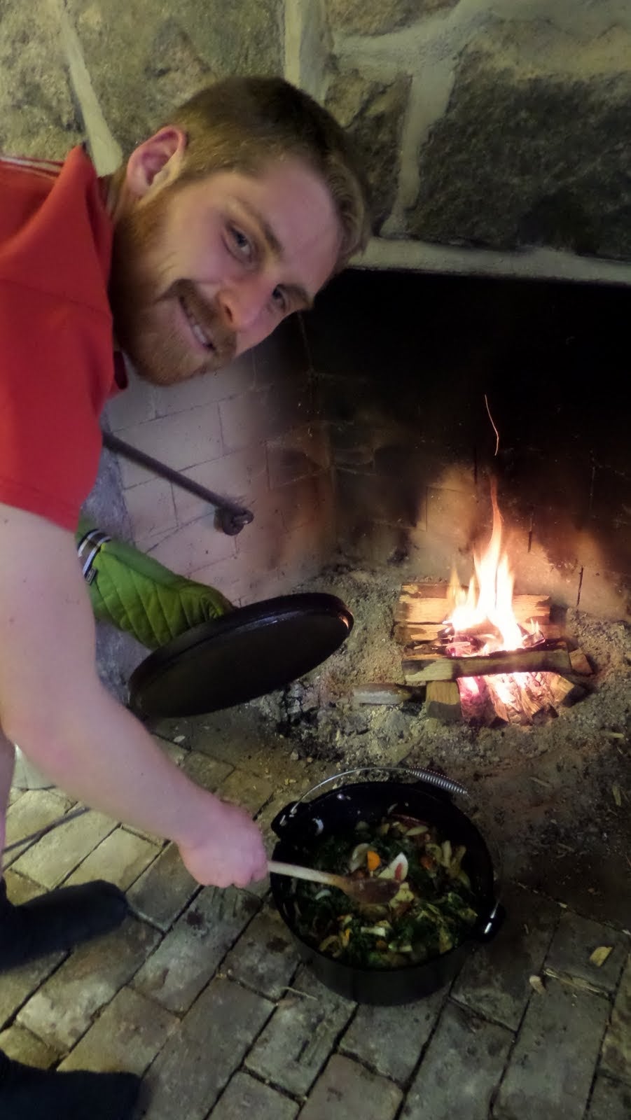 Josh stirs a cast iron pot with vegetables, cooking in the lit stone fireplace.