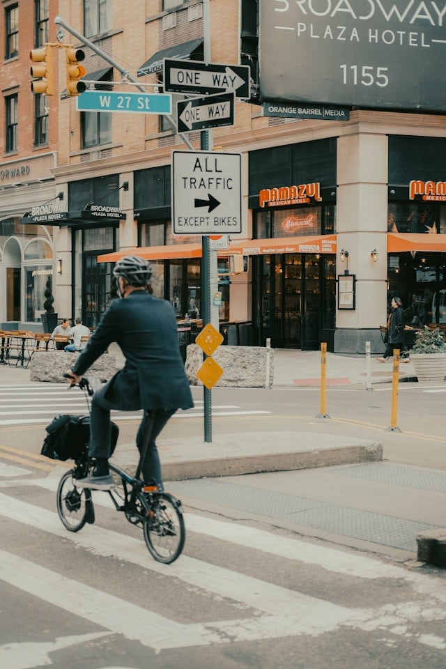 A person in a suit rides a bicycle across a crosswalk near a restaurant with signage, under traffic signals and street signs indicating one-way traffic.