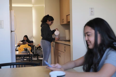 3 students enjoying their residence space. One person is sitting at the kitchen table, while one makes tea in the kitchen, and the last is reading in the living room.