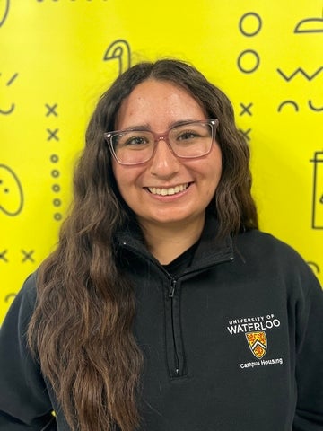 Person in a black UWaterloo branded sweater, standing in front of yellow banner with black branded UWaterloo logos.