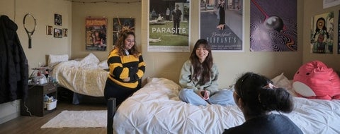 3 women in a Claudette Millar Hall double room talking. One women is sitting on the bed, another standing to her left, and then one in front of her.