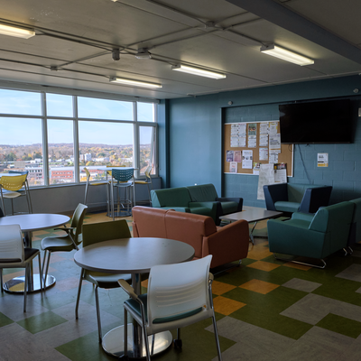 CMH communal floor kitchen and lounge, with a TV mounted on the wall, a bulletin board, several windows on the left, and multiple tables and chairs around the room.