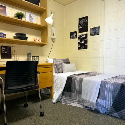 A staged single dorm room with grey plaid bedding and a wooden desk with a chair and shelves above it for storage.