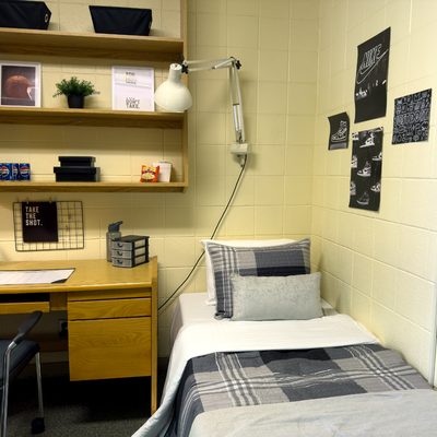 A staged single dorm room with grey plaid bedding and a wooden desk with a chair and shelves above it for storage.