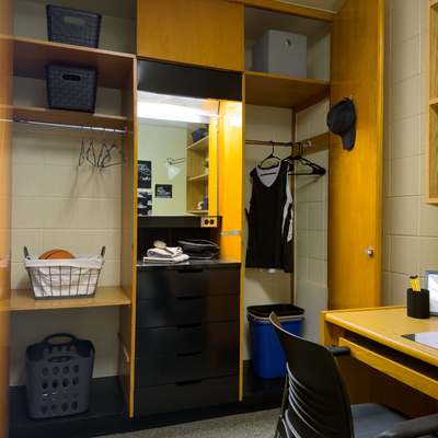 A wooden closet with a mirror and drawers staged with shirts hung up and baskets showing storage space in a single dorm room.