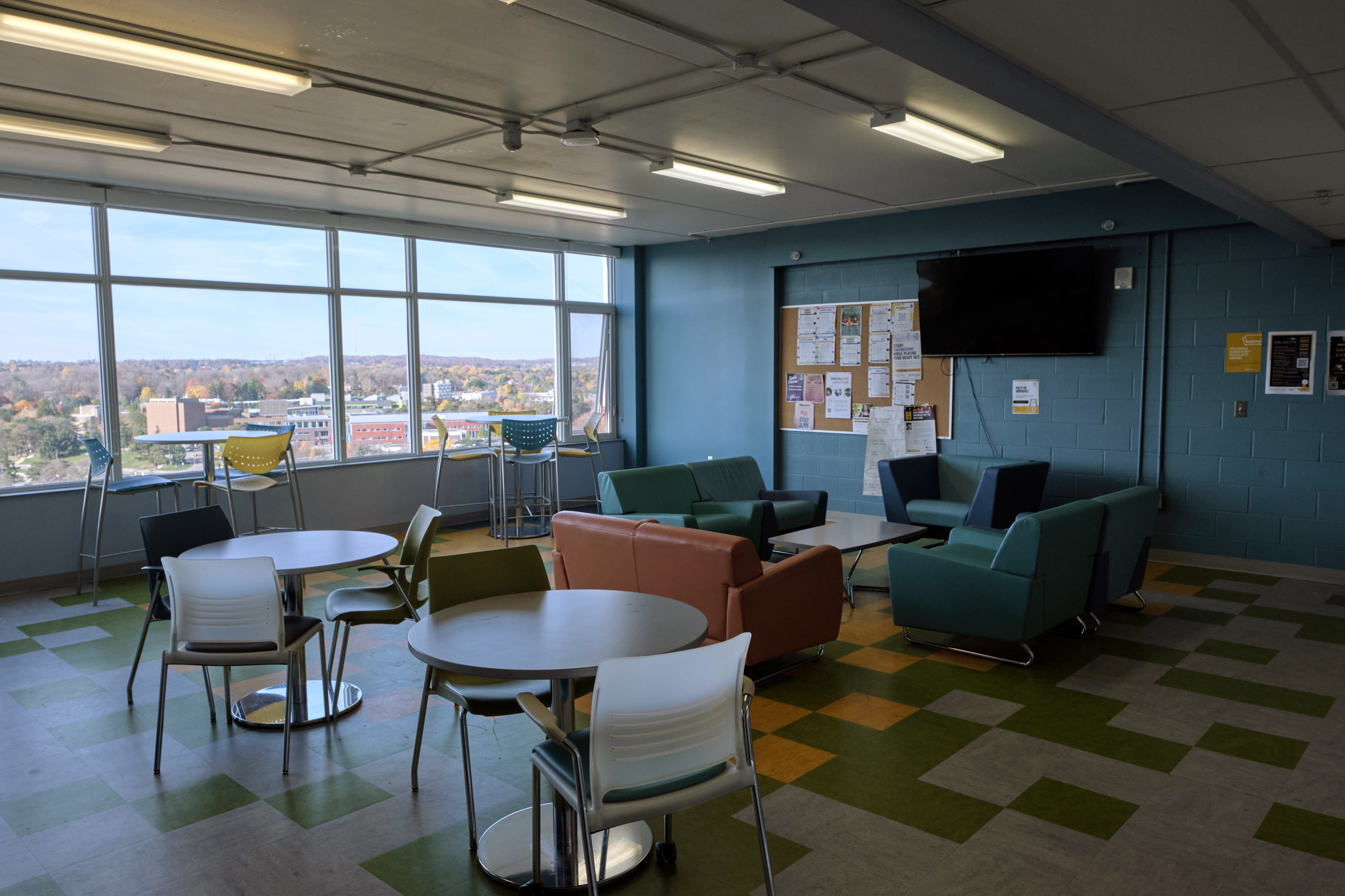 CMH communal floor kitchen and lounge, with a TV mounted on the wall, a bulletin board, several windows on the left, and multiple tables and chairs around the room.