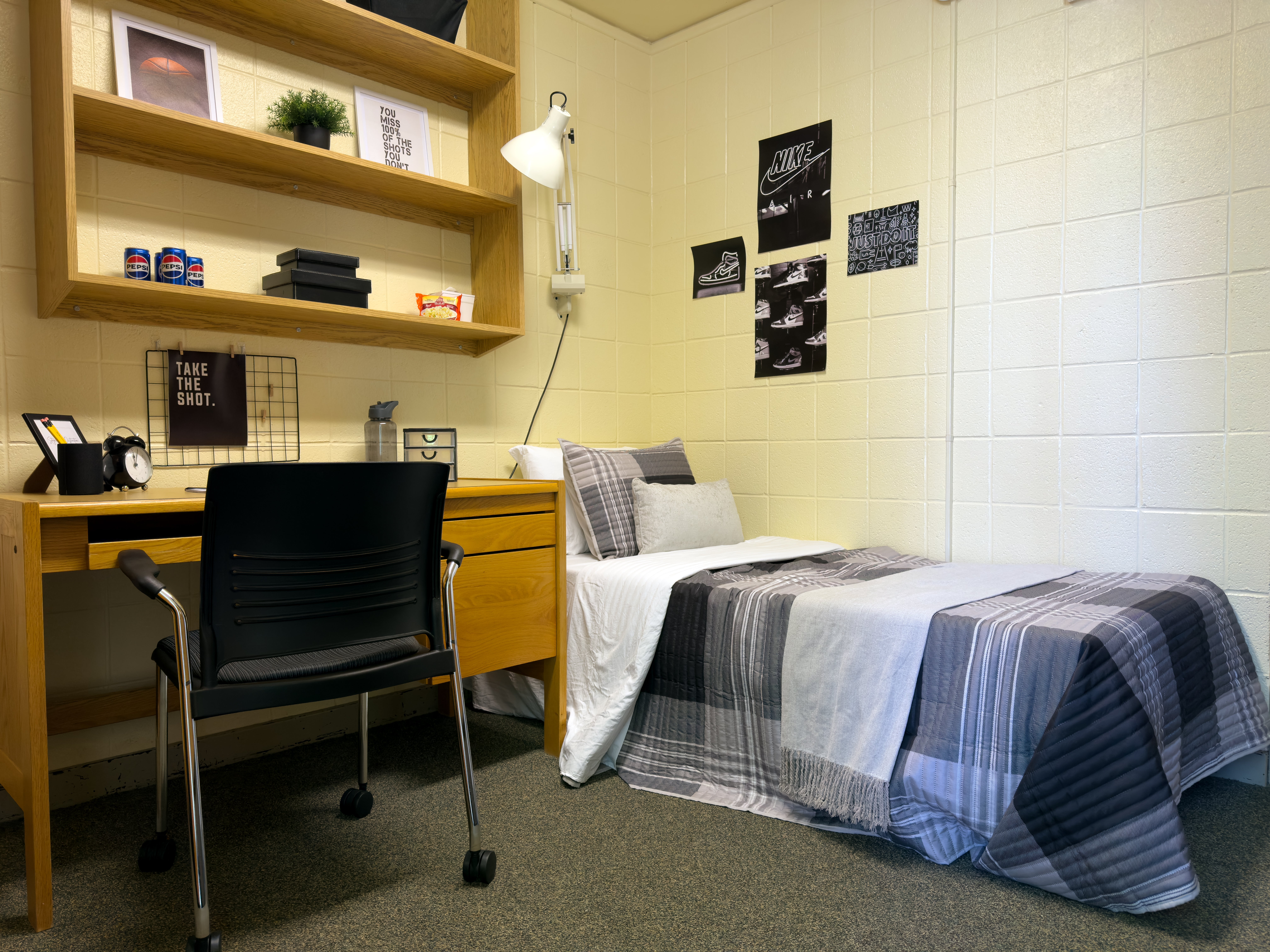 A staged single dorm room with grey plaid bedding and a wooden desk with a chair and shelves above it for storage.
