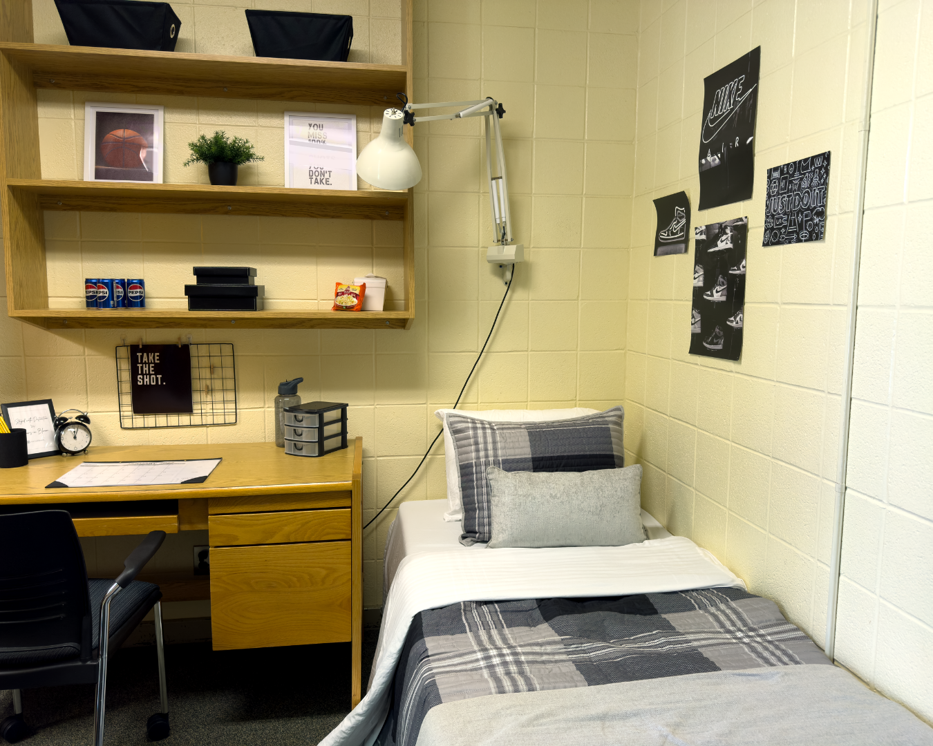 A staged single dorm room with grey plaid bedding and a wooden desk with a chair and shelves above it for storage.
