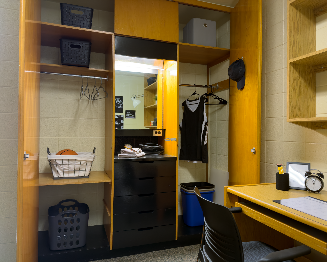A wooden closet with a mirror and drawers staged with shirts hung up and baskets showing storage space in a single dorm room.
