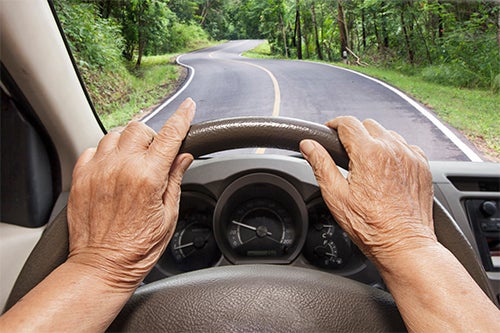 elderly hands on steering wheel driving down road.
