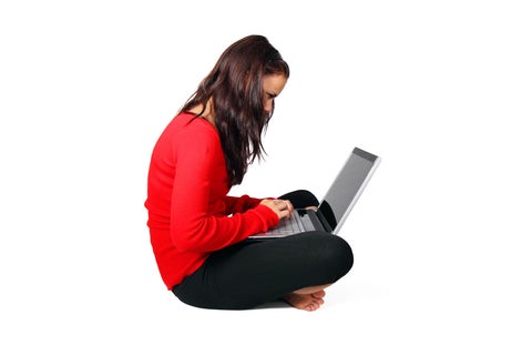 Profile view of young woman in a red shirt and black pants sitting on the floor, with a laptop in her lap.