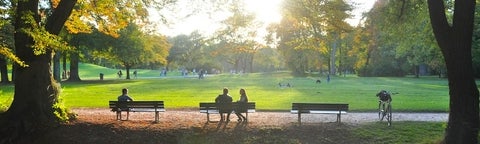 A green park with three benches. One person sitting on the far left. A couple on the centre bench. The sun is shining. There is a bike.