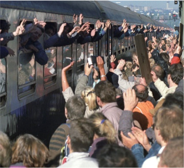 Train with lots of people waving out of partially closed windows. Many people on train platform waving.