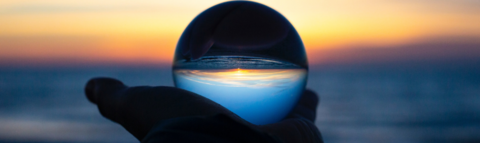 A hand holding a crystal ball through which we see the horizon at sunset. The sky is orange