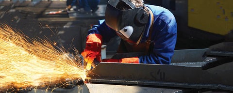Worker wearing welding helmet and gloves welds metal while sparks fly.