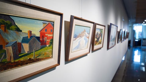 A white wall with a series of framed paintings, farm landscapes in bright colours