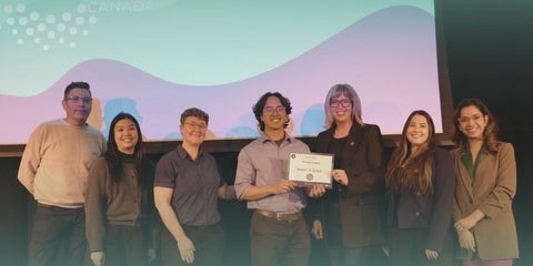Gabriel next to siz other people posing in front of the stage while holding his prize 