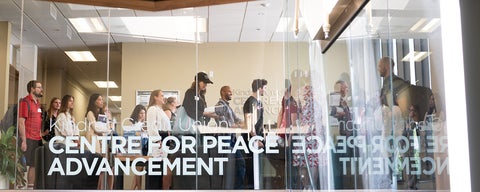 A view from across the front windows of the 4th floor, which houses the Centre for Peace Advancement. A crowd of students on a tour stand in the lobby behind the windows. 
