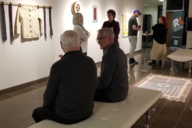 two men on a bench in the center of a gallery with four others standing and chatting behind them