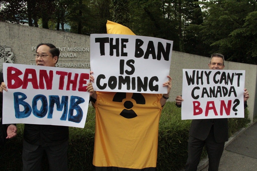 Three people holding up signs with text reading "ban the bombs", "the ban is coming", and  "why can't Canada ban?".