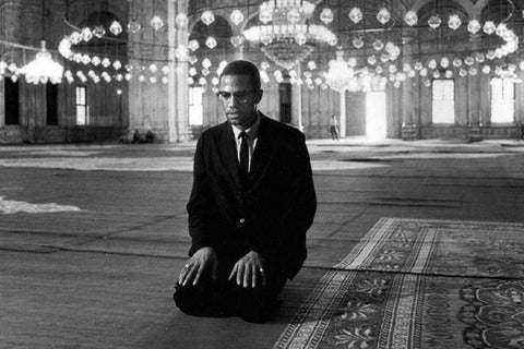 Malcolm X kneels on the ground while wearing a black suit during his 1964 pilgrimage to Mecca