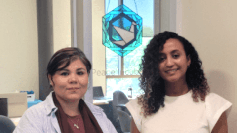 Wazhma and Addi stand in front of the Grebel Peace Incubator dove stained glass window