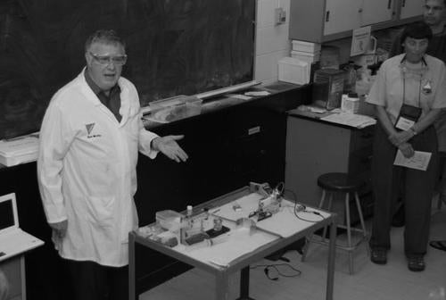 Teacher in lab coat standing behind metal tray with chemistry equipment.