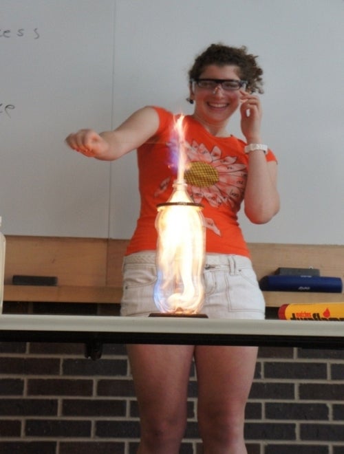 Girl standing behind soda bottle on table containing flame.