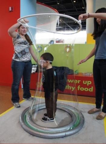 Boy standing inside cylindrical bubble made by two women.