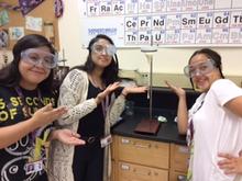 Three girls in a lab pointing to a drop of white material coming out of a funnel. The funnel is supported by an iron ring on a retort stand. There is a periodic table in the background 