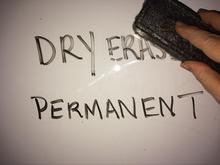 A white board with the words “dry erase” and “permanent” written on it. There is a hand holding an eraser about to try and erase words
