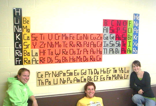 Three people posing under LEGO periodic table on wall.