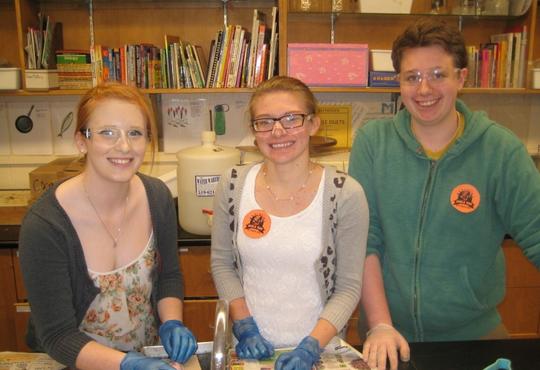 three students smiling in a lab all with goggles 