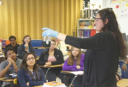 a women in front of a classroom holding up a beaker with a dark blue solution 